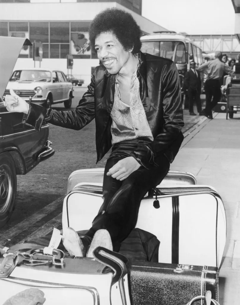 Jimi Hendrix photo from the early '70s. Jimi is sitting on a suitcase and holding his thumb out like he's hitchhiking.