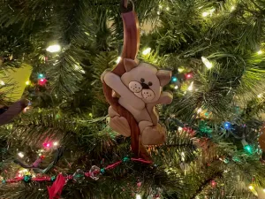 A close up of a holiday ornament on a Christmas tree.. it a cat that looks like it's hanging from a ribon. The ornament is made of wood using the craft style of Intarsia.