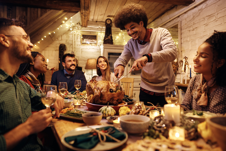 Multiracial group of friends gathering at dining table for Thanksgiving dinner. Focus is on man carving roast turkey.