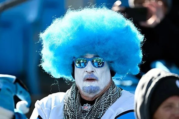 A Detroit Lions fam is wearing a blue wig in the stands at a game. The man also has his face painted white and blue facial hair. This picture is used in an article talking about Detroit Lions must haves.